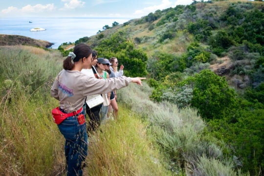 a guide points across a grassy ravine while leading a group of students on a hike through a hilly area with ocean in the background