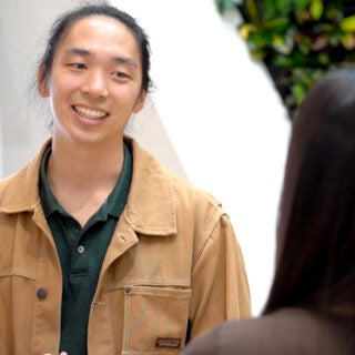 Forrest Lee, wearing a tan jacket and black polo shirt, talks with a coworker in front of a wall of green plants