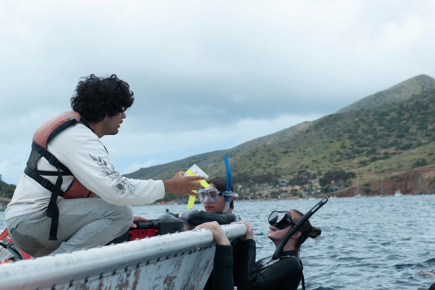 Gabe Romero sits in a boat wearing a lifejacket while passing “clod cards,” or degradable plaster blocks, to Declan Bulwa, who is in the water in snorkeling gear