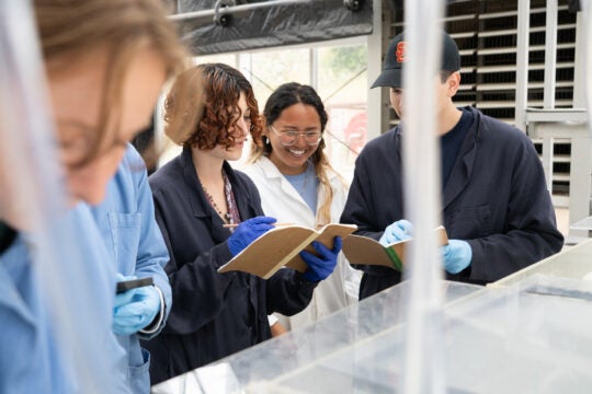 a small group of students wearing lab coats and blue nitrile gloves record data in their notebooks as they stand at a clear plastic tank holding ocean specimens