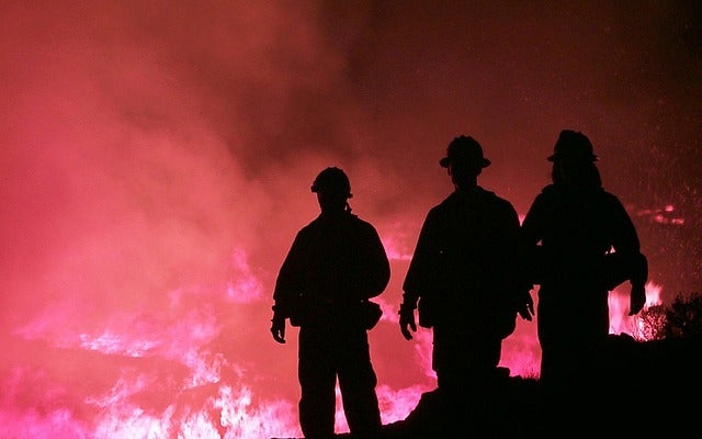 three firefighters stand silhouetted against a raging wildfire