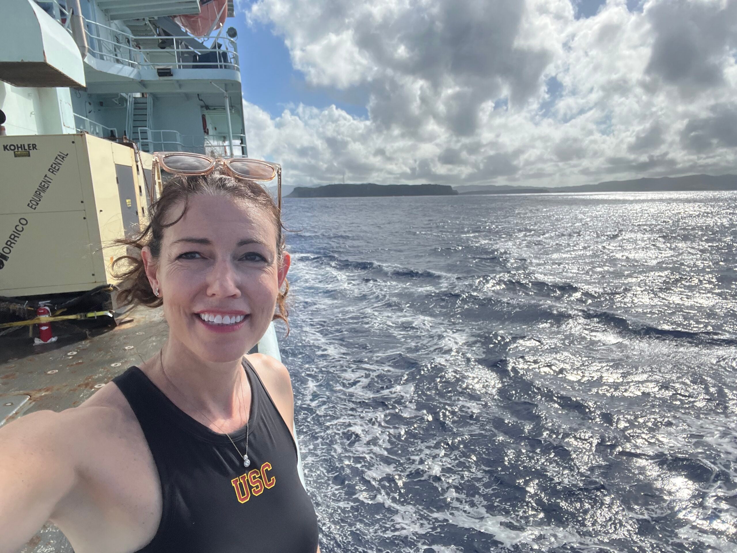 Karen Lloyd, wearing a black tank top with a USC logo, stands on the deck of a ship with ocean and clouds in the background