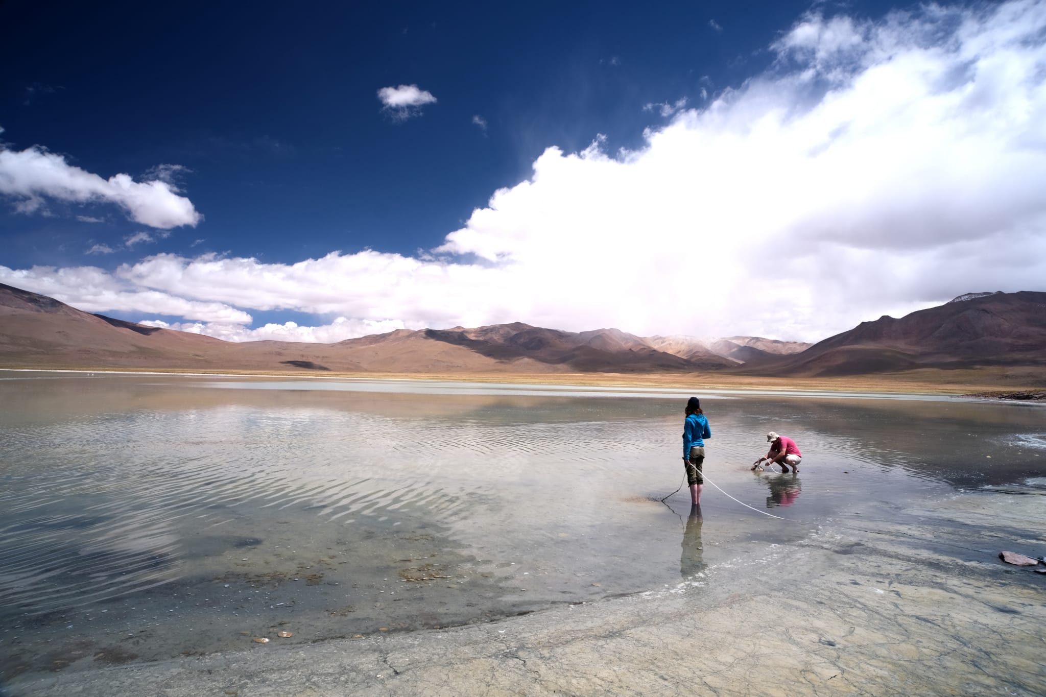 two researchers working in a shallow, clear pond look small against a vast landscape of gray rock and brown hills