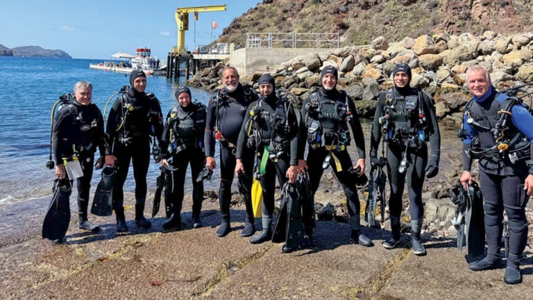 a group of eight NASA divers, dressed in wetsuits and scuba gear, stands on a concrete path just before entering the water