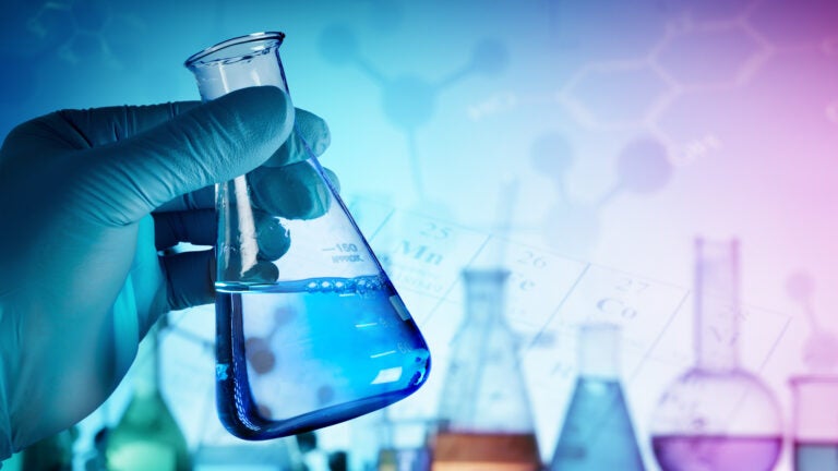 a gloved hand holds a glass beaker of blue liquid in front of a line of lab glassware