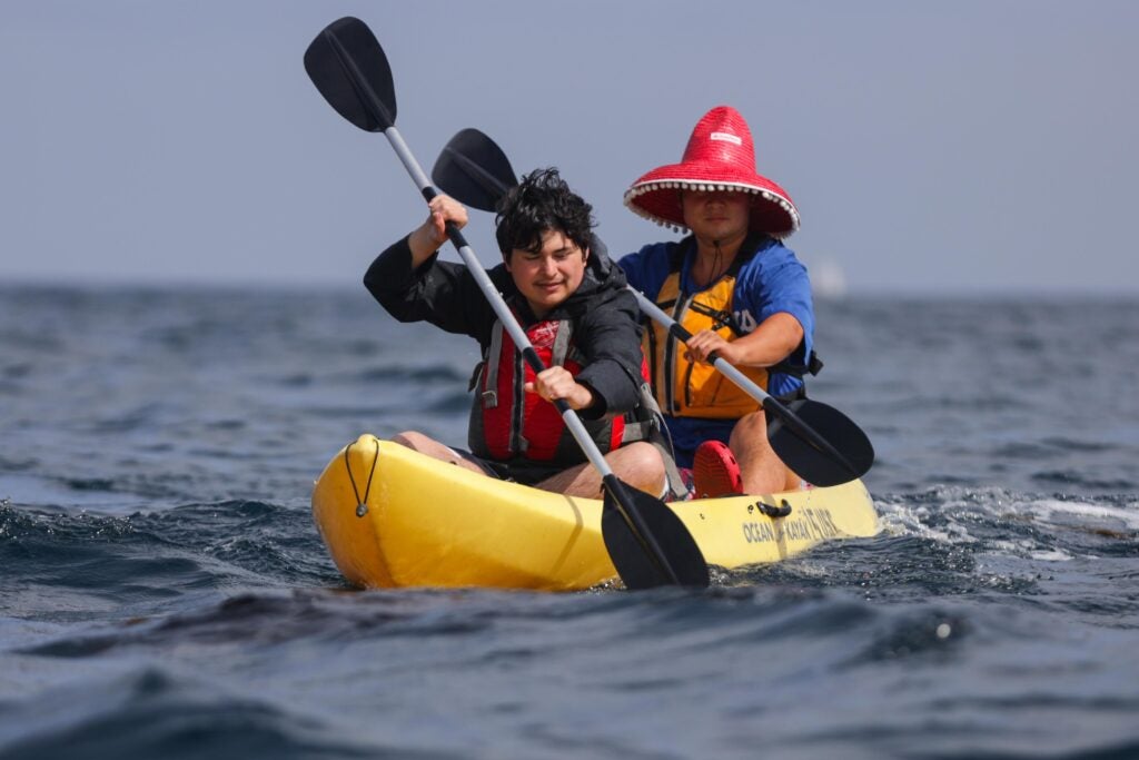 two students, one wearing a red hat, paddle a yellow fiberglass kayak