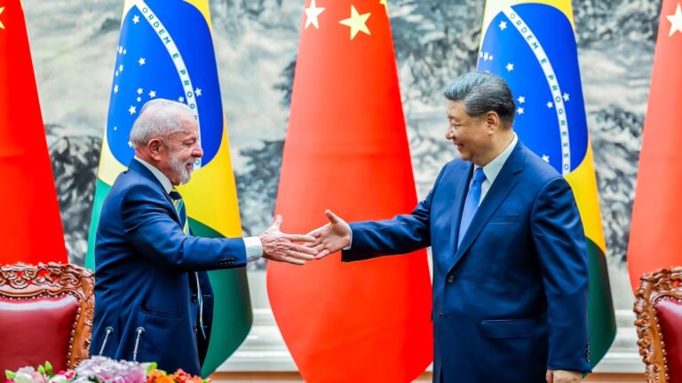 two dignitaries in blue suites shake hands in front of a display of flags