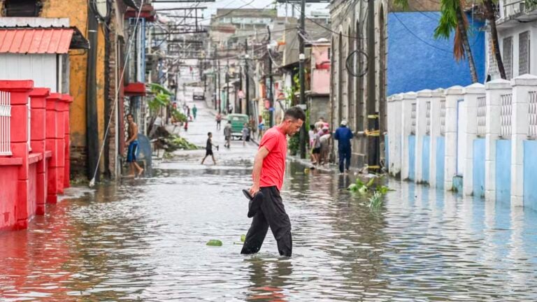 a person in a red shirt and black pants holds their shoes while wading across a flooded street in the Caribbean