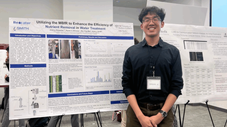 a smiling student wearing a black buttondown shirt and olive green pants stands in front of a research poster at a symposium