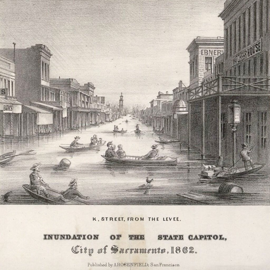 a historic, black-and-white lithograph shows people rowing in small boats down a street lined by wooden buildings in 1800s Sacramento