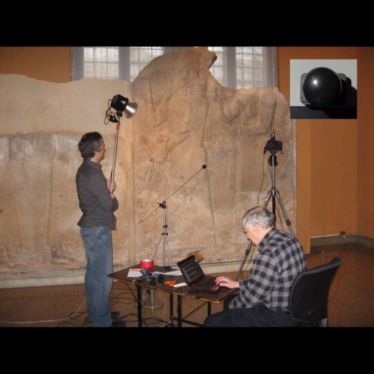 One man standing with flash, second man seated with laptop computer. A large inscribed stone is in the background. Illustration.